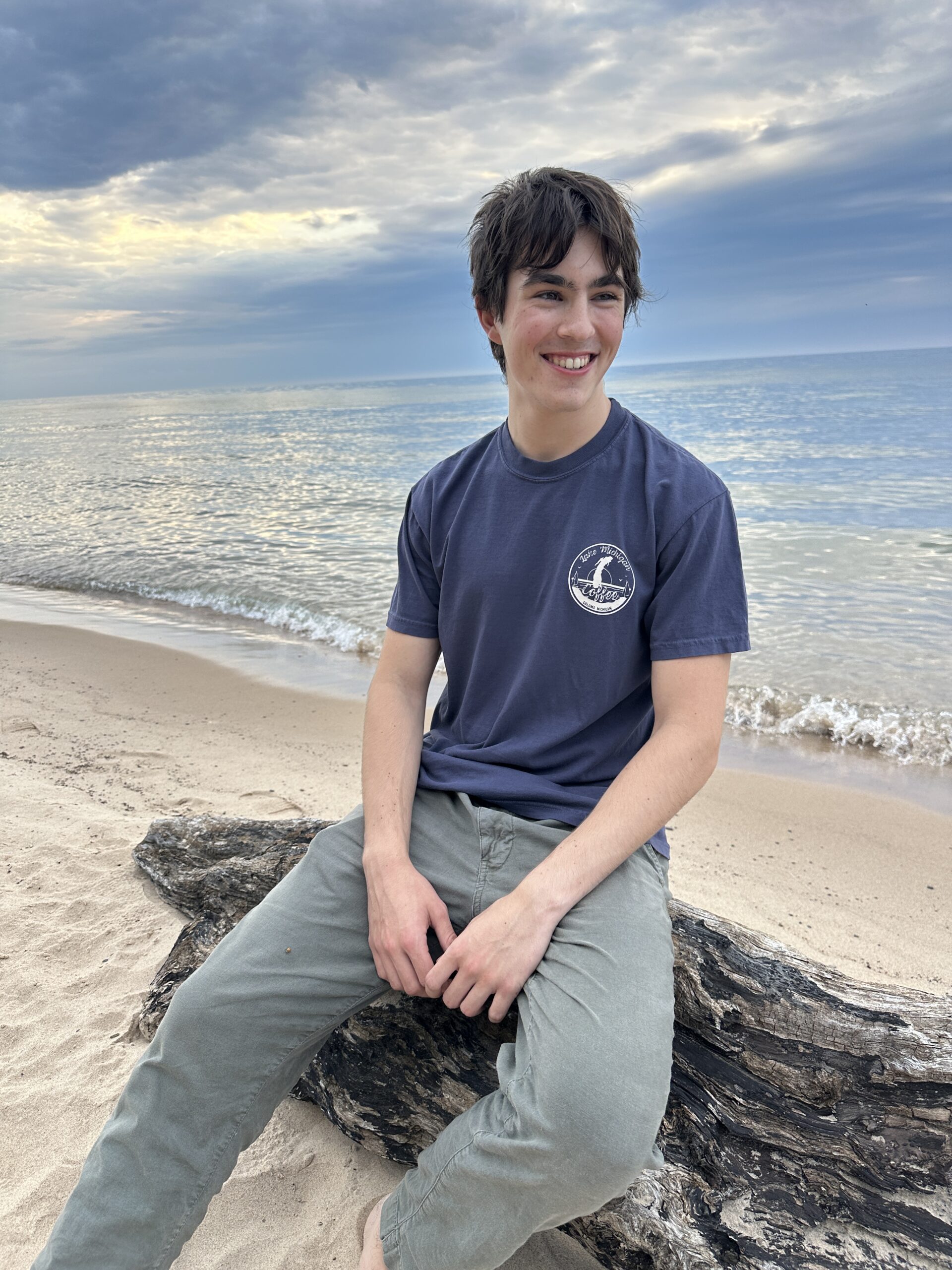 Home A man in a navy Lake Michigan Coffee tee sitting on driftwood by the lake